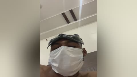 A video of a young Black man wearing a surgical mask and a blue surgical cap, taken from a low angle, showing his upper body in a sterile, white-walled room with recessed ceiling lights.