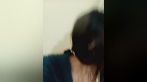 A blurry video of a person with dark hair and a teal jacket, partially visible in a dimly lit room with a beige wall.