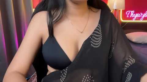 Video of a light-skinned woman with long black hair, wearing a black, sheer blouse with a floral pattern and a black bra, sitting in a dimly lit bedroom with a neon sign reading \"Tighty\" in the background.