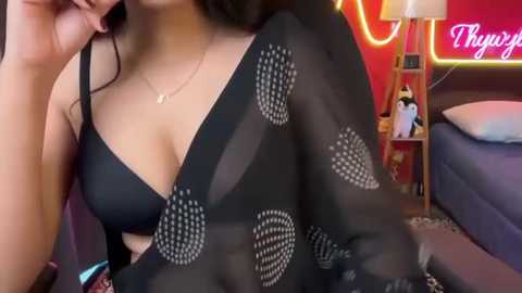 Video of a woman in a black bra with a sheer, patterned overlay, leaning on a bed with a pillow. Background features a red wall with neon lights and a stuffed animal.