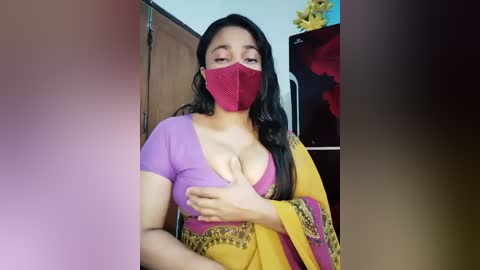 Video of a South Asian woman with long black hair, wearing a purple blouse and yellow sari with red mask, covering her cleavage, in a dimly lit room with yellow flowers.