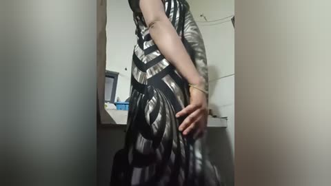 Video of a person in a black and white striped dress, standing in a dimly lit room with white walls. The person's right hand is raised, and their left hand is on their hip.