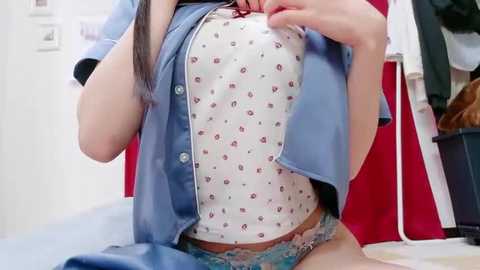 Video of a light-skinned Asian woman with long black hair, lifting a blue button-up shirt to reveal a white bra with red hearts. She's wearing blue lace panties. Background shows a red and white room with a hanging jacket.