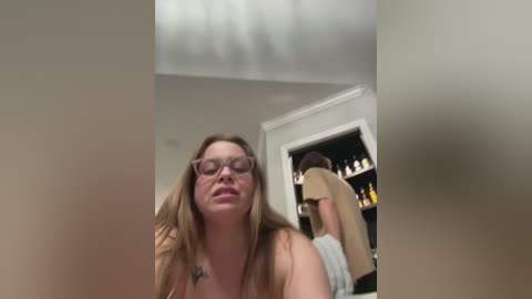 A video captures a young woman with glasses, fair skin, and long brown hair, looking towards the camera. Behind her, a man in a beige jacket stands in a dimly lit room with shelves of bottles.