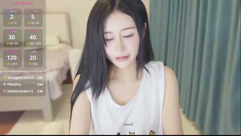 Video of an Asian woman with long black hair, wearing a white sleeveless top, looking down, in a modern bedroom with blue curtains and a bed.