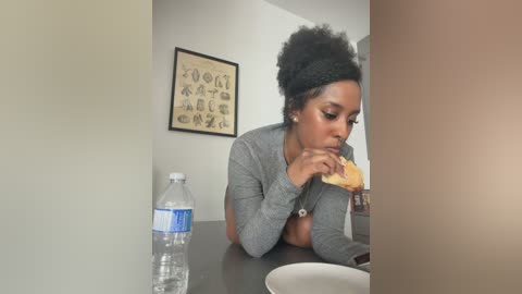 Video of a young Black woman with a large afro, wearing a gray cardigan, eating a sandwich at a table in a minimalist room. Water bottle and framed artwork in background.