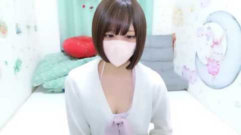 Video of an Asian woman with short, dark hair, wearing a white mask, light pink blouse, and white cardigan in a pastel-themed room with a bed, green pillows, and a red heart pillow.