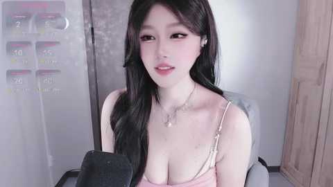 Video of an East Asian woman with long black hair, fair skin, and large breasts in a low-cut pink top, sitting in a dimly lit room with a white door and a digital display on the left.
