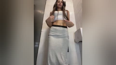 Video of a woman in a white tank top and skirt, looking down the tiled hallway, with a paper towel dispenser on the right.