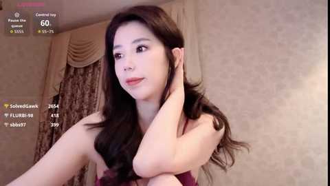 Video of a young East Asian woman with long, dark hair, wearing a sleeveless maroon dress, posing in a dimly lit room with beige floral wallpaper and heavy curtains.