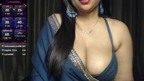 A video of a woman with long black hair, wearing a blue, low-cut blouse with intricate gold embroidery, and a silver necklace. The background features a dark curtain and a digital clock displaying 20:00.