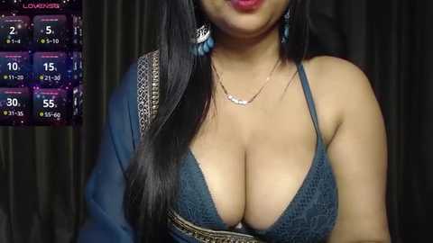 Video of a woman with medium brown skin, long black hair, wearing a dark blue crochet bralette that accentuates her large breasts, with a silver necklace and blue earrings. Background shows a virtual reality interface with weather and game stats.