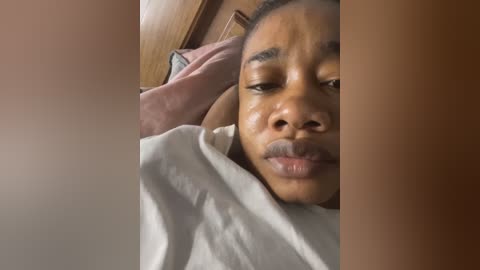 A close-up video of a young Black woman with a medium skin tone and short hair, lying on a pink pillow with a white blanket covering her chest. The background shows a wooden headboard.