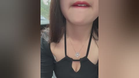Video of a young woman with light skin, dark hair, and red lipstick, wearing a black halter top with a floral pendant necklace. The background is blurred.