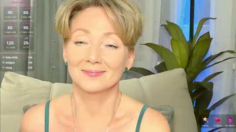 Video of a middle-aged woman with short blonde hair, wearing a teal tank top, smiling, sitting on a light grey couch, in a room with a green plant in the background.