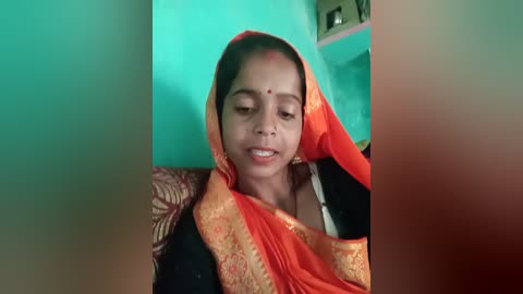 Video of a young South Asian girl with medium brown skin, wearing a bright orange and gold sari, sitting indoors against a teal wall. She has a serene expression, with a red bindi on her forehead.