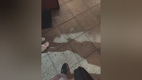 Video of a man's feet in black shoes on a beige tiled floor, shadow cast by a person unseen, partially visible legs, and a brown object (chair?) in the background.