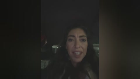 Video of a woman with curly black hair, smiling broadly, wearing a black leather jacket, set in a dimly lit room with blurred background objects.