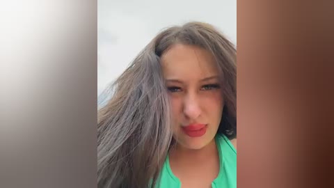 Video of a young woman with long, straight, dark brown hair, fair skin, and bright red lipstick, wearing a green top. Background is blurred, featuring a brown wall on the right and a white wall on the left.