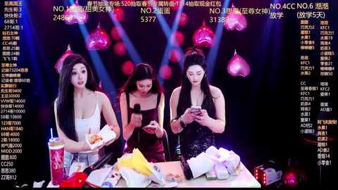 Video of four women, one Asian, one Caucasian, and two East Asian, sitting at a table, holding smartphones, surrounded by food and drinks, under dim, blue-lit stage lights.