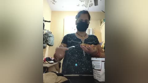 Media: Video of a woman in a dark floral shirt, black mask, and bracelets, holding a bowl and spoon, standing in a cluttered kitchen with beige walls and hanging utensils.