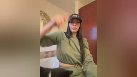 Video of a young woman with long black hair, wearing a green striped top, black cap, and green pants, standing in a dimly lit room with beige walls and a red door.