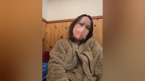 Video of a woman with a light complexion, wearing a beige, fuzzy jacket, a black mask covering her nose and mouth, and brown hair, sitting on a wooden bed with a blue blanket, in a cozy, rustic room.