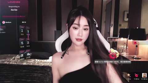 A video of a young East Asian woman with long black hair and pale skin, wearing a black off-shoulder dress, sitting in a dimly-lit bedroom with a TV and bed visible in the background.