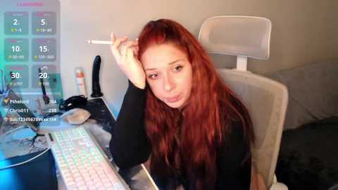 Video of a young woman with long, wavy red hair, wearing black clothing, sitting at a computer desk, holding a pen, in a modern, cluttered room.