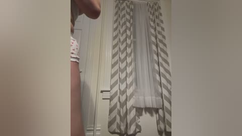 Video of a woman with light skin, wearing a white shirt with red polka dots, standing in a bathroom with a herringbone-patterned shower curtain.