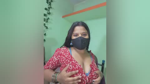 Video of a woman with medium skin tone and straight black hair, wearing a red patterned blouse, black mask, and bracelets. She stands in a simple room with green walls, a leafy plant, and a window.