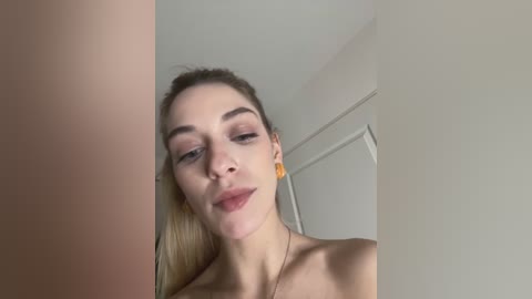 A close-up video of a young Caucasian woman with fair skin, straight blonde hair, and minimal makeup, wearing large yellow earrings. She's topless, with a neutral expression. The background shows a plain, white room with a door.