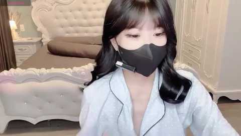 A video of an Asian woman with long black hair, wearing a black face mask and light blue robe, sitting in a plush, white tufted bed with white wooden nightstands and drawers.