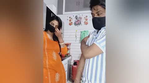Video of two South Asian women in a kitchen; one in a bright orange sari, the other in a striped shirt, both masked, chatting.