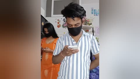 Video of a young man in a striped blue and white shirt, black face mask, and orange shirt, looking at his phone in a cluttered room with a woman in the background.