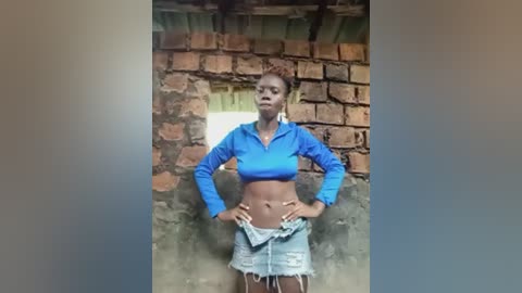 Video of a young Black woman with braids, wearing a blue crop top and denim shorts, standing against a rustic brick wall with a window.