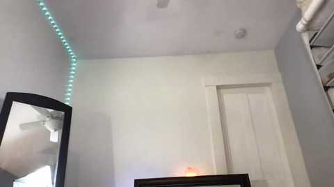 A video of a modern, minimalist bedroom with white walls, a black mirror, a ceiling fan, and a teal string light. A white door and closet are visible in the background.