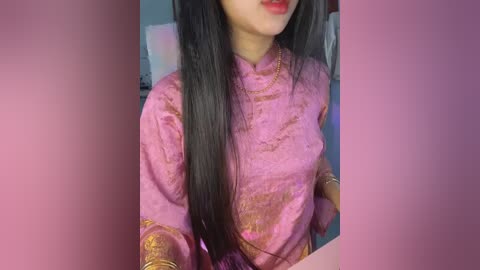 Media: Video of an Asian woman with long black hair, wearing a pink traditional silk blouse with gold embroidery, and red lipstick, in a soft-focus background.