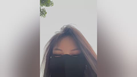A close-up video of a person with long, dark hair, partially obscured by a black face mask. The background is blurred, suggesting an indoor setting with soft, diffused lighting. The image has a slight vignette effect, emphasizing the subject.