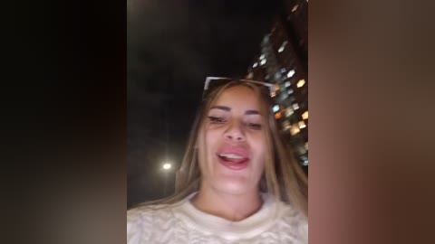 Video of a smiling young woman with long blonde hair, wearing a white knit sweater and dark sunglasses, standing in front of a lit-up, blurred background.