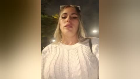 A video of a blonde woman in a white blouse, looking relaxed with closed eyes and a slight smile, taken at night under dim lighting.