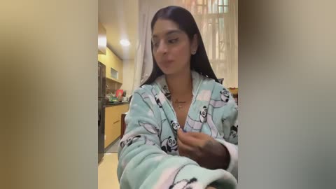 Video of a young woman with long black hair, wearing a light blue robe with cartoon characters, sitting in a modern kitchen with yellow walls and white cabinets.