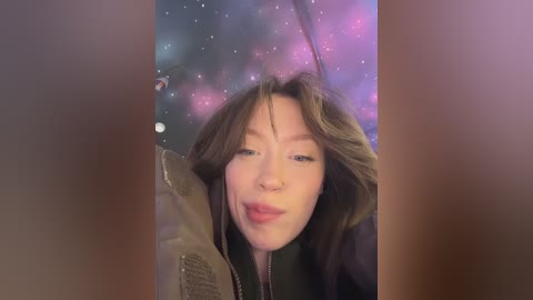 A video of a young woman with long, wavy brown hair, wearing a green jacket, smiling with her lips slightly parted, set against a vibrant, cosmic background with stars and planets.