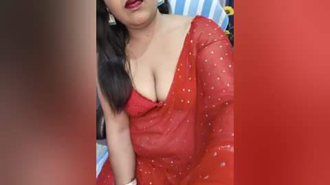 A video shows a South Asian woman with medium skin tone, black hair, and red lipstick, wearing a red saree with a gold blouse, partially revealing her cleavage. The background features blue and red stripes.