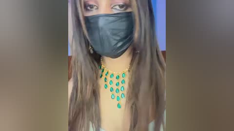 A video of a person with long brown hair, wearing a black face mask, a turquoise necklace, and a green dress. The background is blurred, and the lighting is soft.