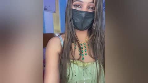 Video of a light-skinned woman with long, straight brown hair, wearing a black face mask, green dress, and gold necklace. Background shows a yellow poster and blue wall.
