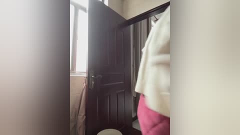 A video of a narrow, dimly lit bathroom doorway, featuring a dark wooden door with a metal handle, and a beige wall. A white towel hangs on the left, and a person wearing a white sweater and red pants is partially visible in the right foreground.