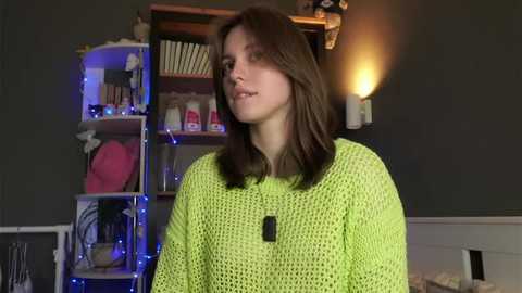 Video of a young woman with straight brown hair, wearing a neon green crochet sweater, standing in a dimly lit room with dark green walls, a wooden bookshelf, and a lit sconce.