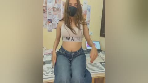 Media: Video of a young woman with medium brown skin, wearing a black face mask, a white sports bra, and blue jeans, sitting on a bed with a laptop and messy room in the background.