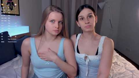 Video of two young Caucasian women with long hair, in casual attire, sitting on a bed with beige sheets, in a dimly lit room with soft lighting.